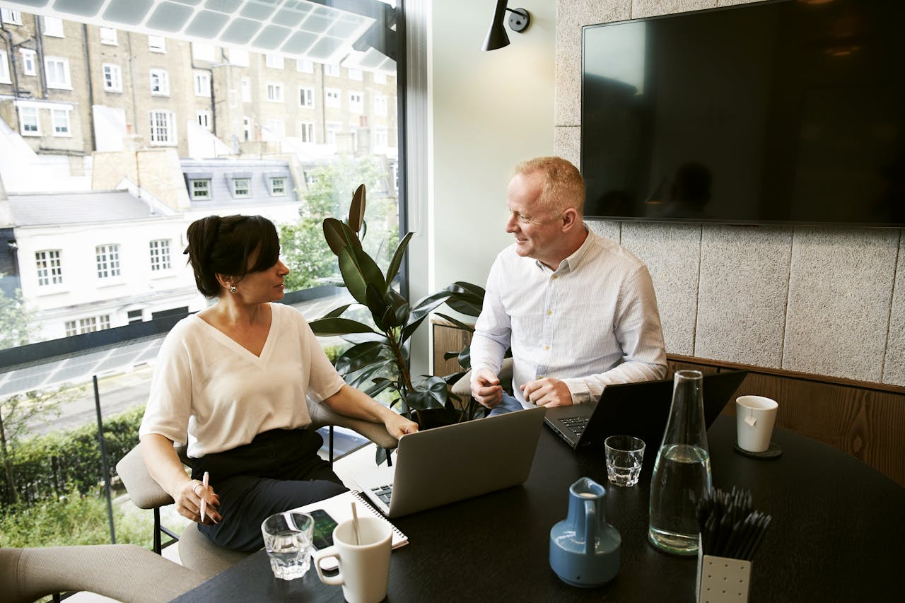 about-us-01 Two business professionals engaging in a team meeting in a modern London office with a large window view.